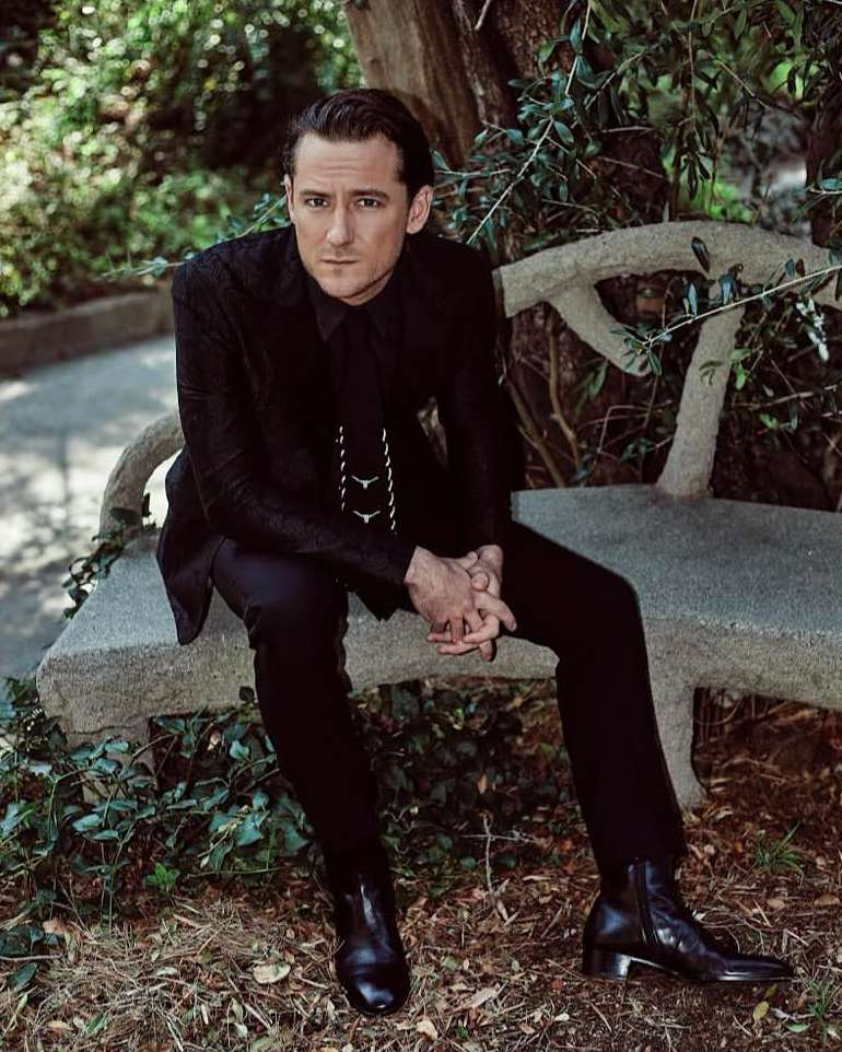Adult male sitting on a stone bench in a garden, wearing a black suit and boots, looking at the camera with a neutral expression.
