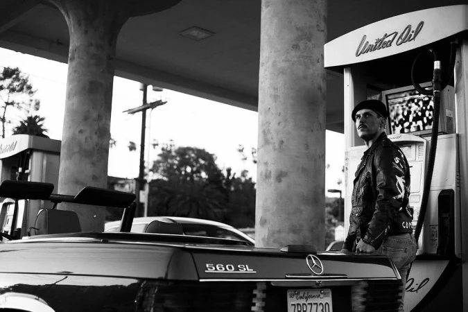 Monochrome image of a man in a leather jacket next to a vintage convertible and gas pump at an outdoor gas station location.
