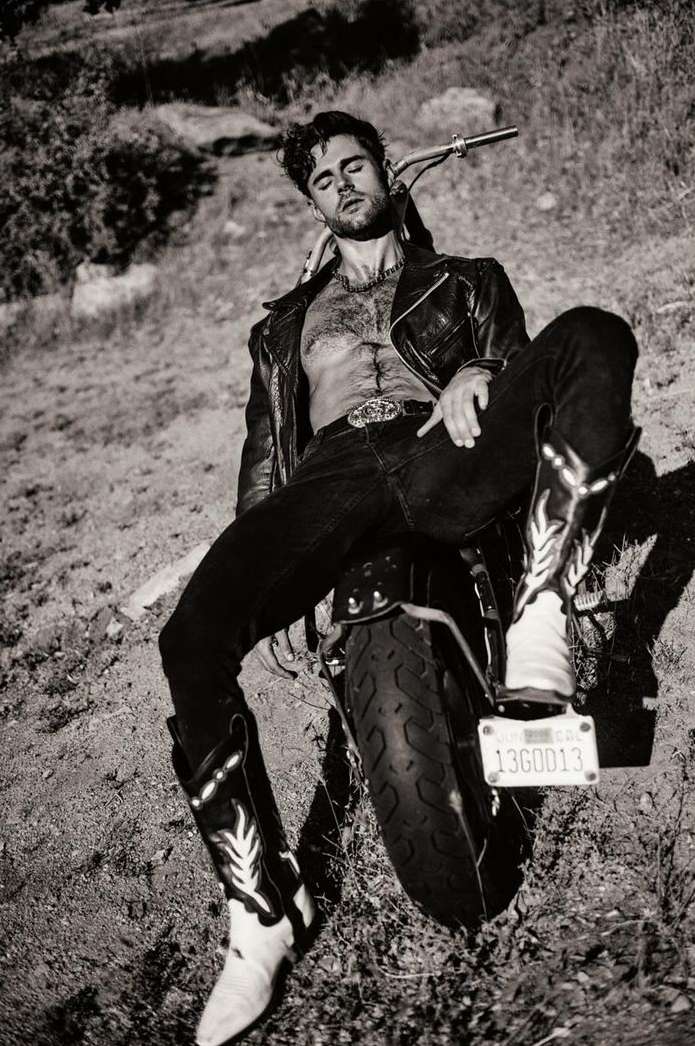 Monochrome portrait of a man in a leather jacket and cowboy boots posing against a motorcycle outdoors.