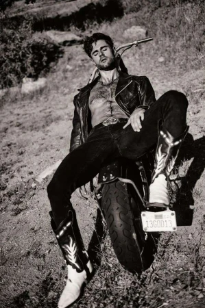 Monochrome portrait of a man in a leather jacket and cowboy boots posing against a motorcycle outdoors.