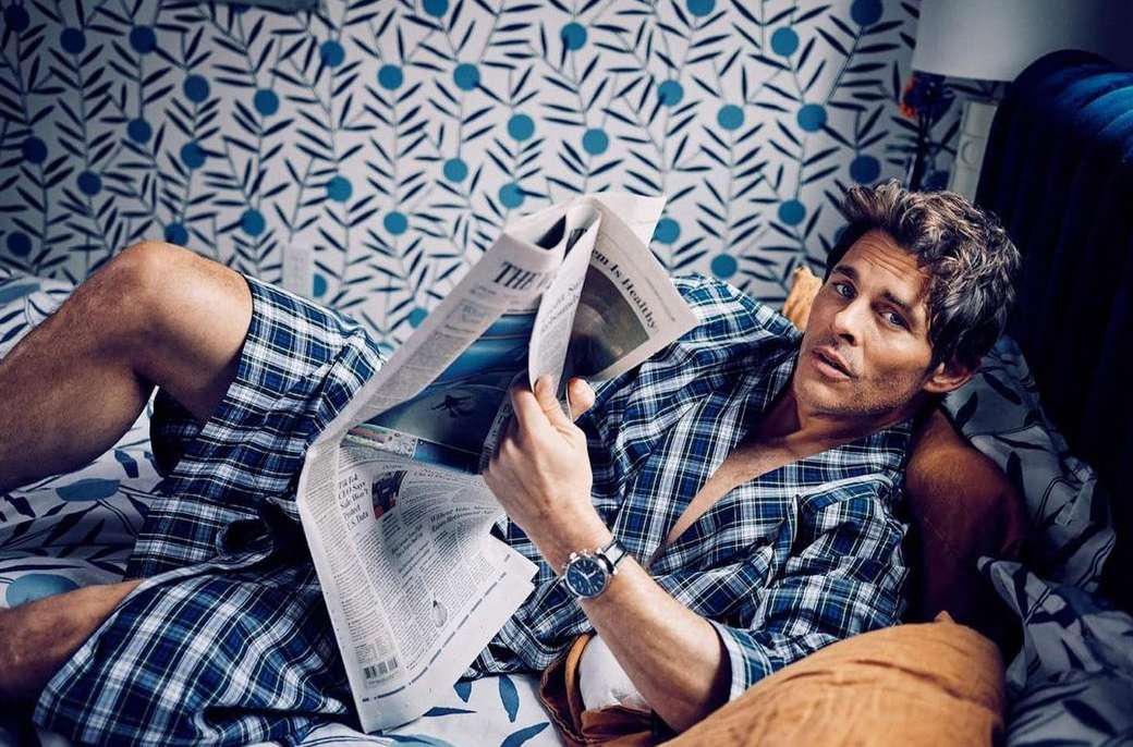 Adult male lying on a bed wearing a plaid robe while reading a newspaper with a blue and white patterned wallpaper in the background.