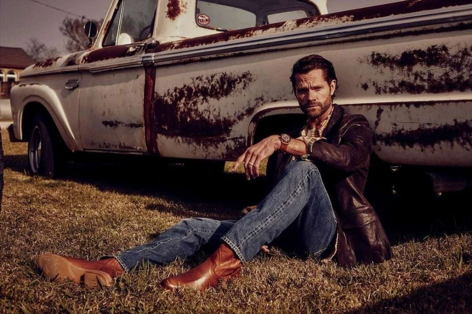 Man sitting on grass beside a rusty pickup truck, wearing a leather jacket and jeans, looking at the camera in an outdoor setting.