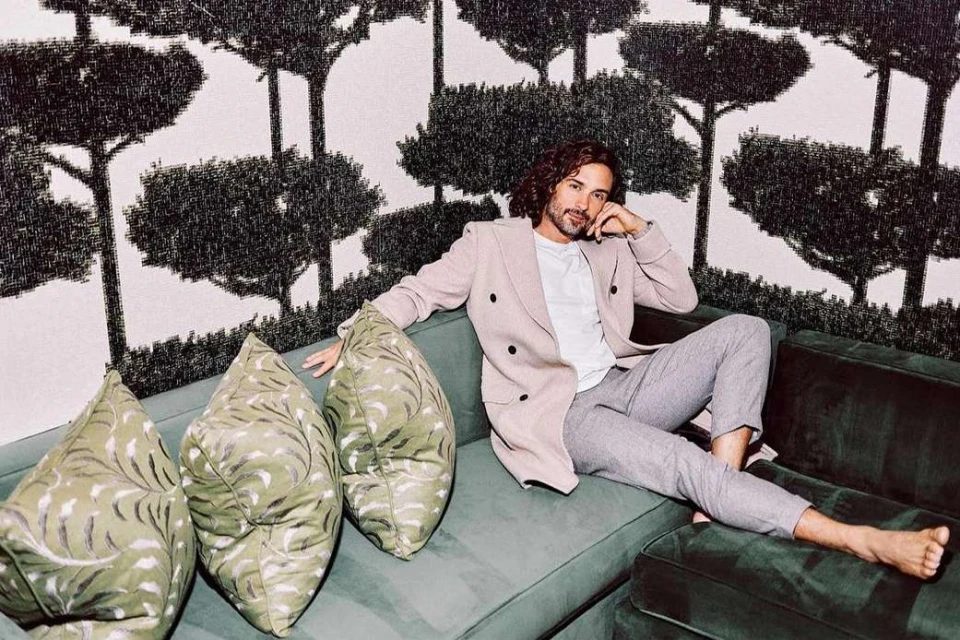 Adult male relaxing on a green couch with decorative pillows against a tree-patterned wallpaper in a casual setting.