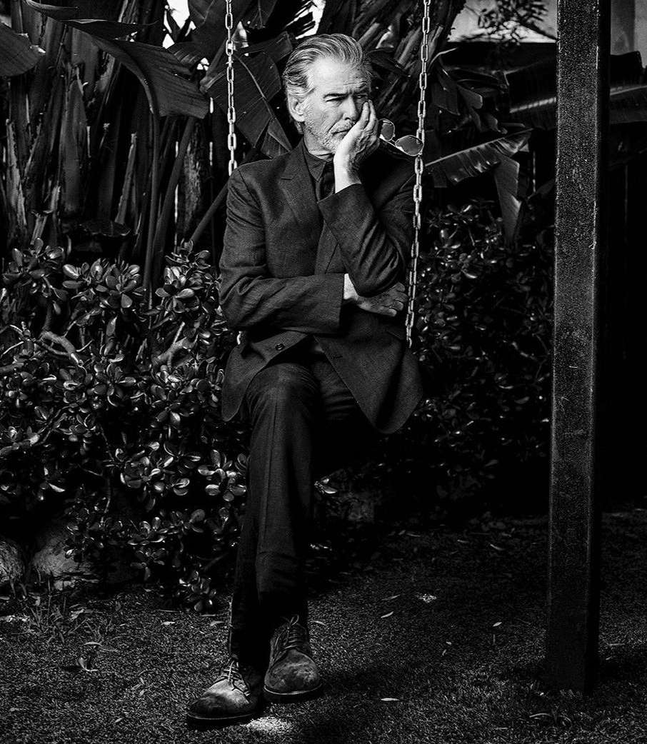 Black and white portrait of a mature adult male in a suit sitting on a swing amidst lush green foliage in an outdoor environment.