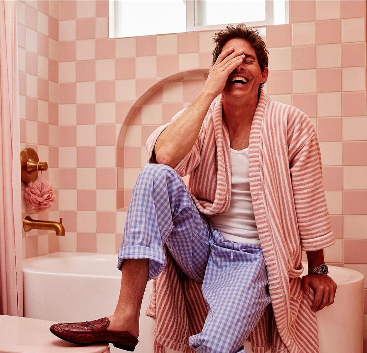 Adult male laughing in a pink bathroom, wearing a bathrobe and pajamas while seated on the edge of a bathtub with tile background.