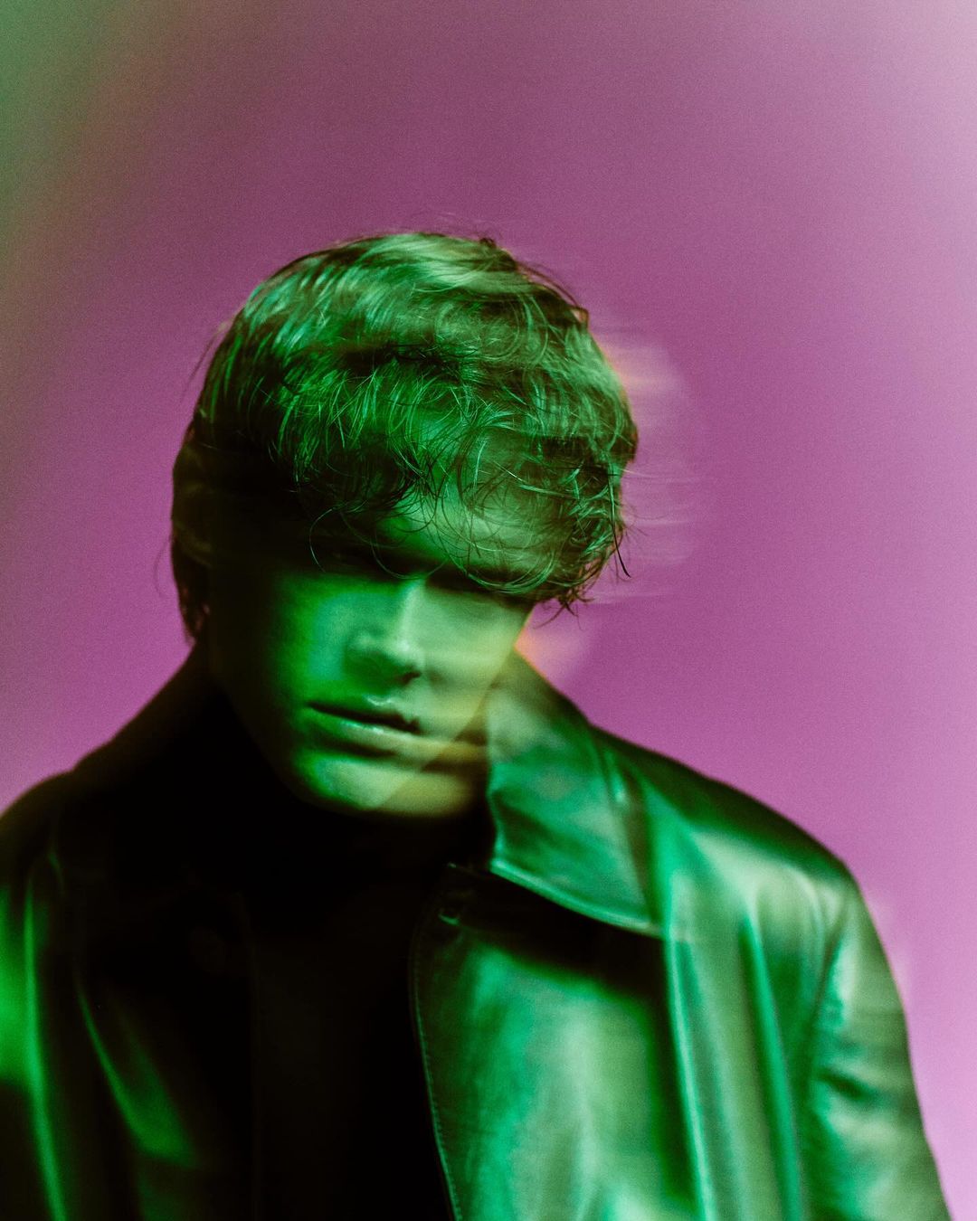 Portrait of a young adult male with light brown hair wearing a black leather jacket, illuminated by green and pink neon lights.