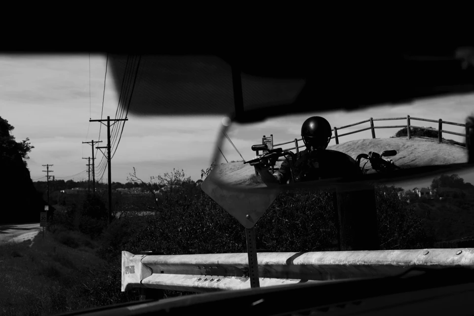 Monochromatic outdoor shot through a car's rearview mirror: a motorcyclist with a helmet on a road against a cloudy sky backdrop.