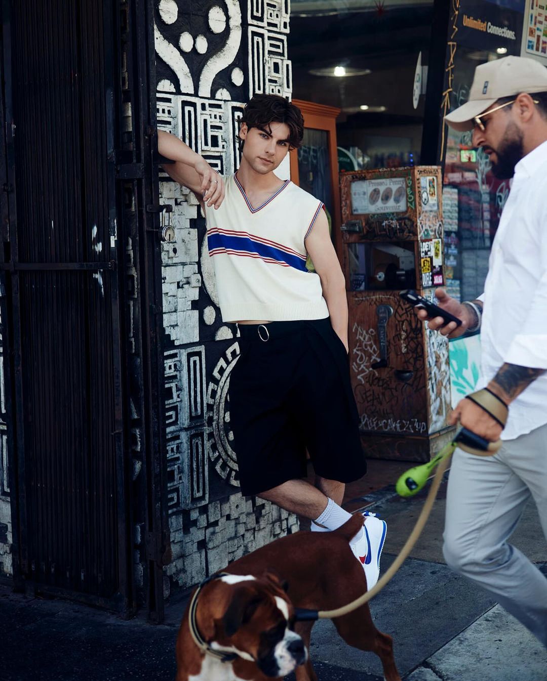 Young man in sleeveless top leans against patterned wall outdoors, Boxer dog, and passerby with phone and hat in a city scene.