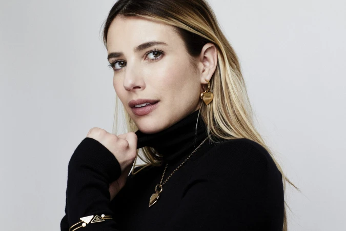 Portrait of Emma Roberts wearing a black turtleneck and gold heart earrings against a white seamless background