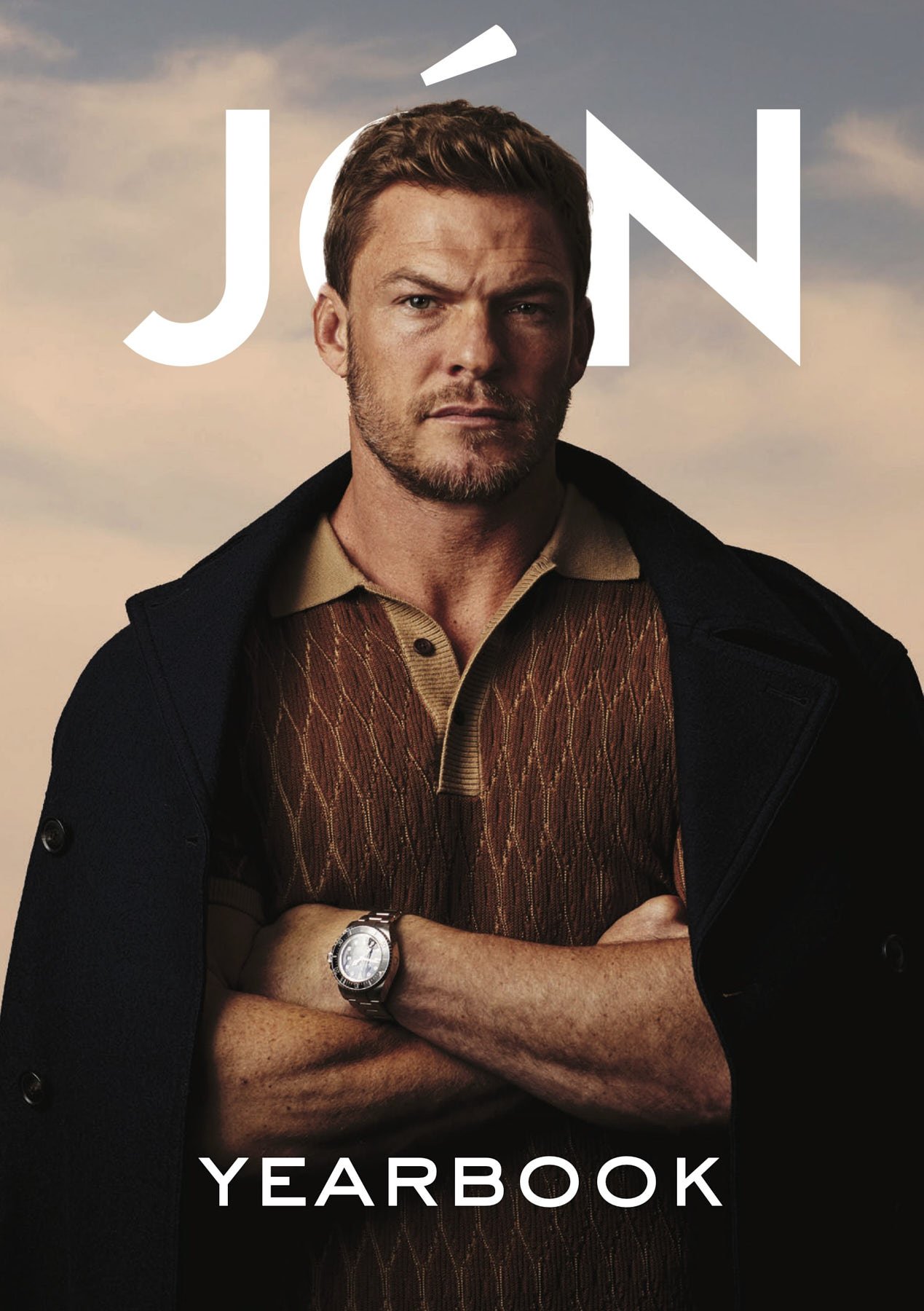 Headshot of actor Alan Ritchson looking directly at the camera with arms crossed wearing a dark coat against a peach background.