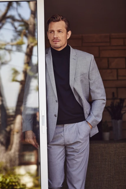 Portrait of a handsome man in a grey suit and black turtleneck standing near a doorway with light reflecting off of the glass.