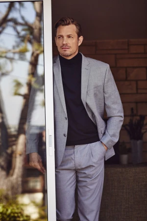 Portrait of a handsome man in a grey suit and black turtleneck standing near a doorway with light reflecting off of the glass.