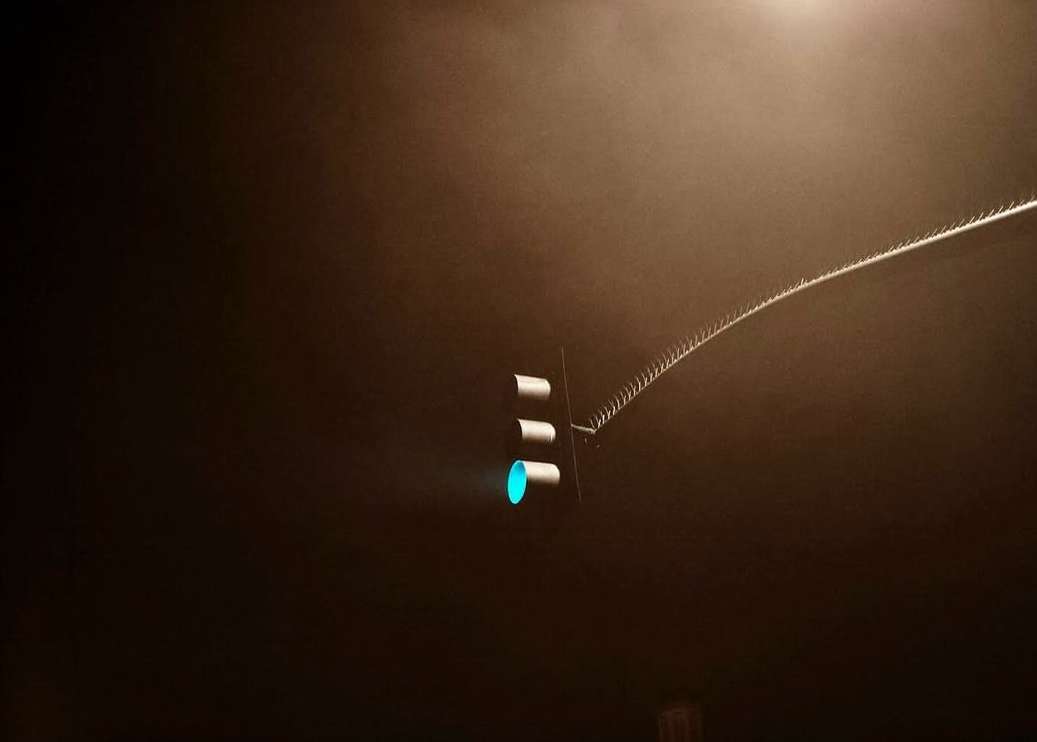 A traffic light glows green in the darkness, attached to a metal arm extending into a hazy night sky, casting a surreal urban scene.