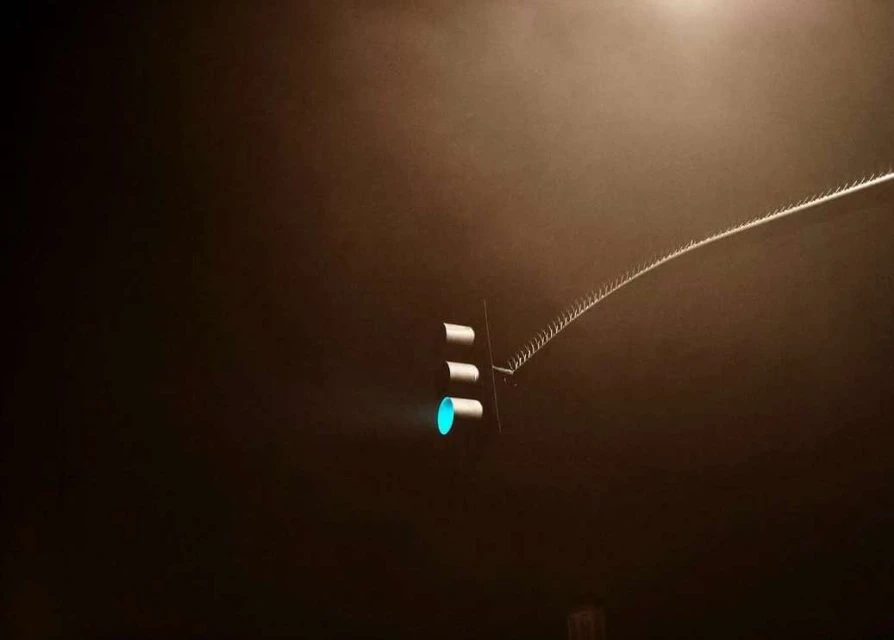 A traffic light glows green in the darkness, attached to a metal arm extending into a hazy night sky, casting a surreal urban scene.