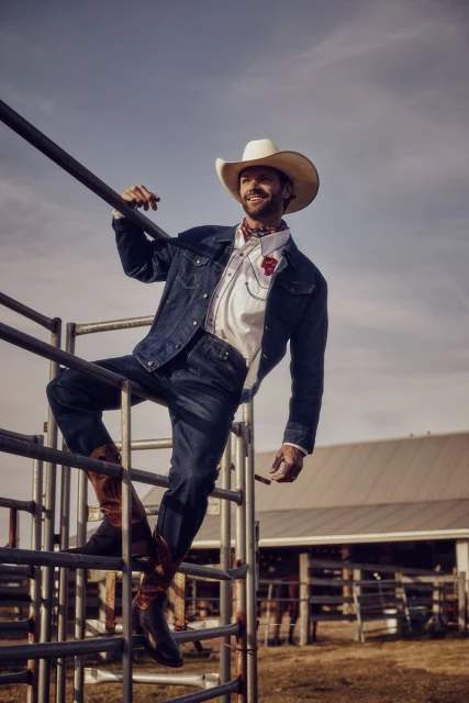 Man in cowboy hat and denim jacket poses on a metal fence with farm building in the background, under a blue sky on sunny day.