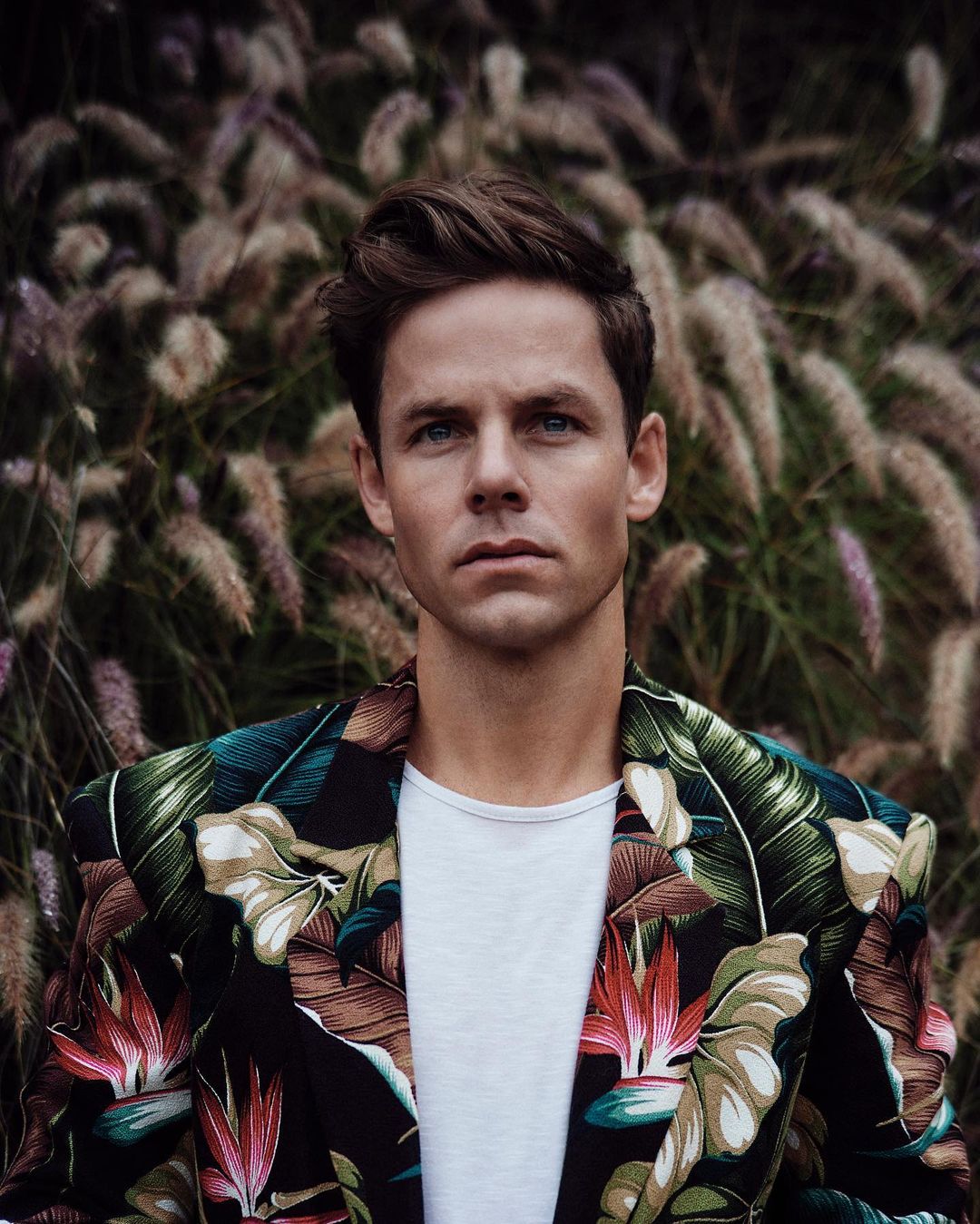 Portrait of a man in a floral-patterned blazer posing outdoors in front of tall ornamental grass with a neutral expression and direct gaze.