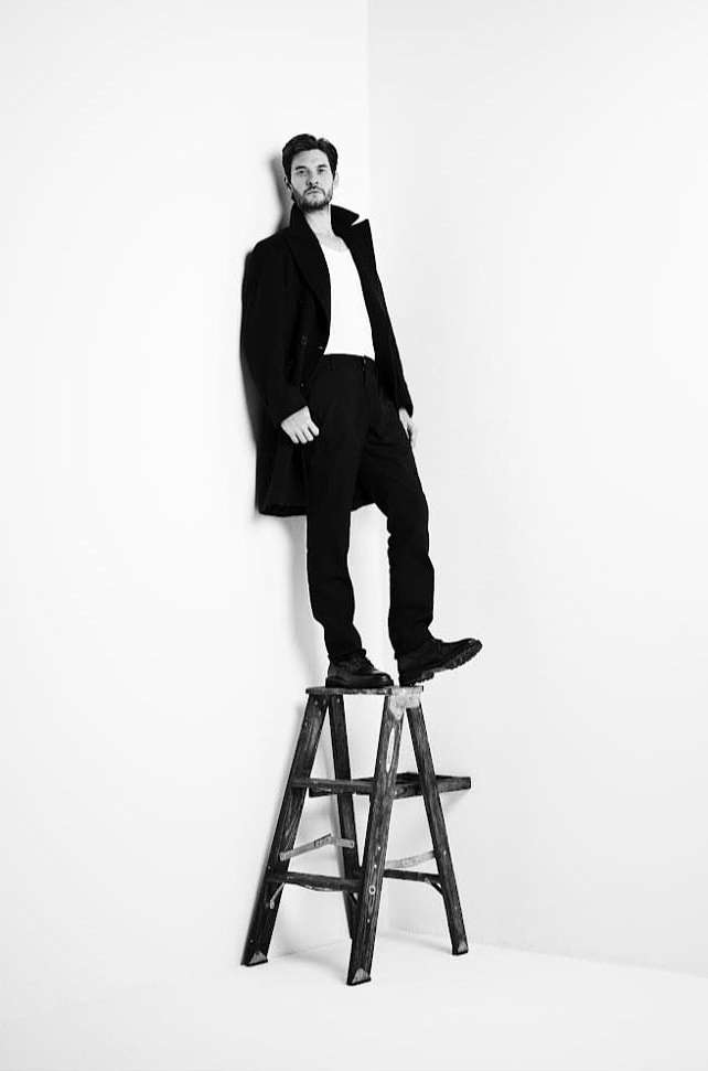 Black and white studio shot of a man in a coat and white shirt standing on a wooden step stool against a white wall backdrop.