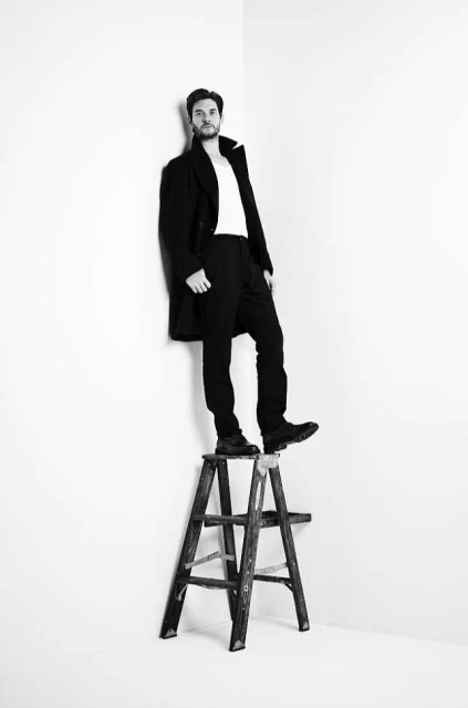Black and white studio shot of a man in a coat and white shirt standing on a wooden step stool against a white wall backdrop.