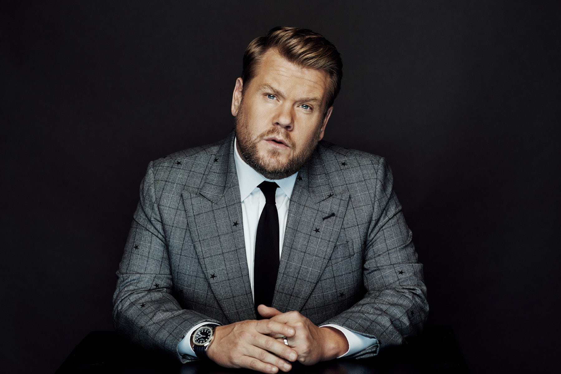 Portrait of a blond adult male wearing a grey suit with black tie, looking at the camera, indoors against a dark background.