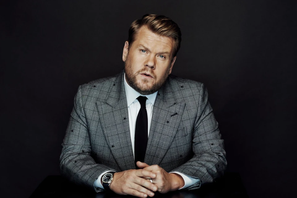 Portrait of a blond adult male wearing a grey suit with black tie, looking at the camera, indoors against a dark background.