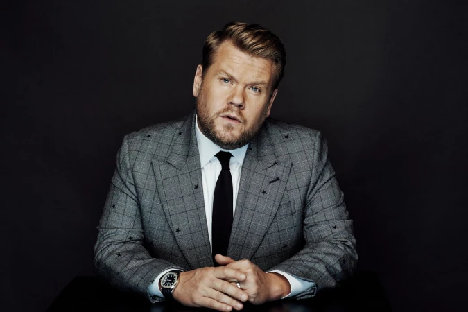 Portrait of a blond adult male wearing a grey suit with black tie, looking at the camera, indoors against a dark background.