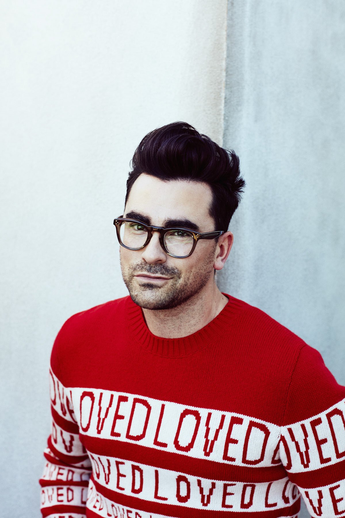 Portrait of a man with dark hair and glasses, wearing a red sweater with white text against a pale blue background.