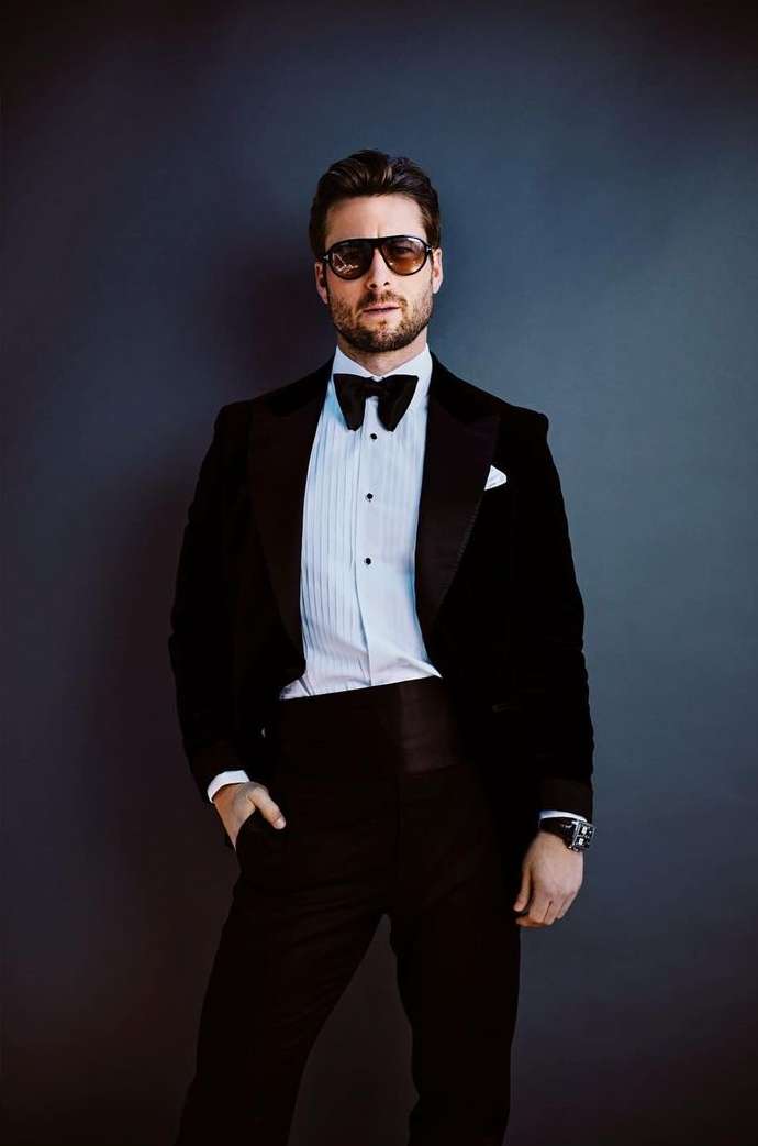 Stylish man in a black velvet tuxedo, bow tie, and sunglasses poses against a dark grey background in a confident portrait.