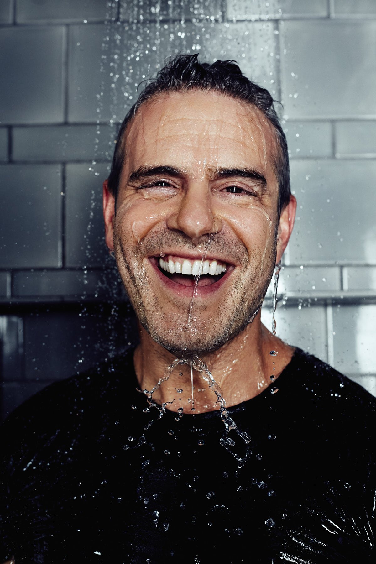 Smiling adult male under shower, wearing a black T-shirt, against gray tiled wall, with water droplets and refreshing vibe.