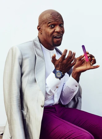 Portrait of Terry Crews wearing a grey jacket and holding a pink lipgloss, posing in front of a white background.