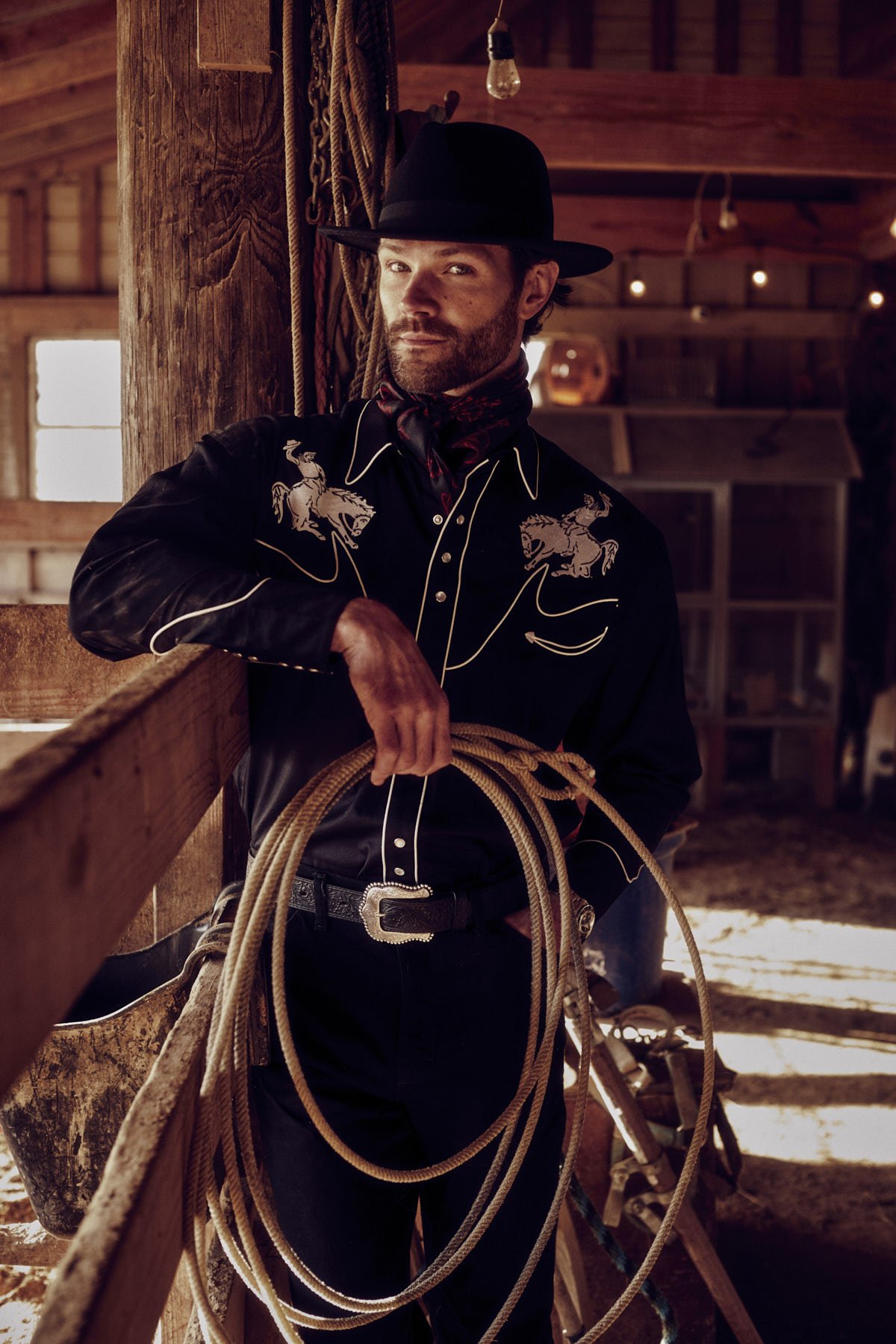 Portrait of an adult male in a black western-style shirt and black hat, holding a lasso, in a wooden barn setting with soft lighting.