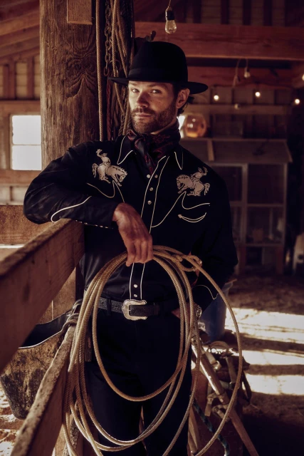 Portrait of an adult male in a black western-style shirt and black hat, holding a lasso, in a wooden barn setting with soft lighting.