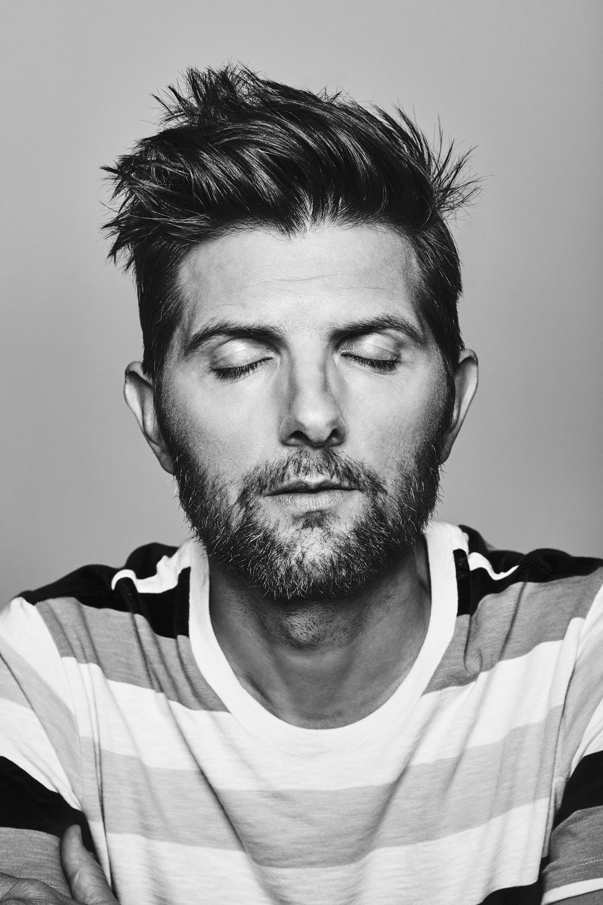 Monochrome portrait of adult male with dark hair and beard, eyes closed, wearing a horizontally striped shirt against a grey background.