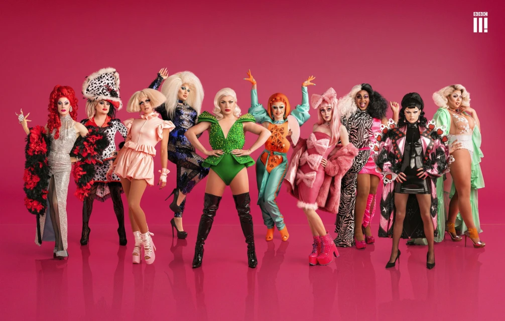 Group of drag queens in vibrant outfits posing against a bright pink backdrop, showcasing diverse styles and expressions, high fashion.