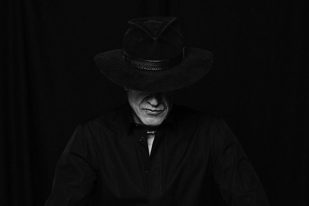 Monochrome portrait of an adult male with a serious expression, wearing a dark cowboy hat and a black shirt against a dark background.