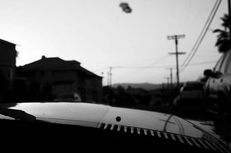 Black and white photo from inside a car, showing the hood, a residential street, utility poles, houses, and distant mountains.