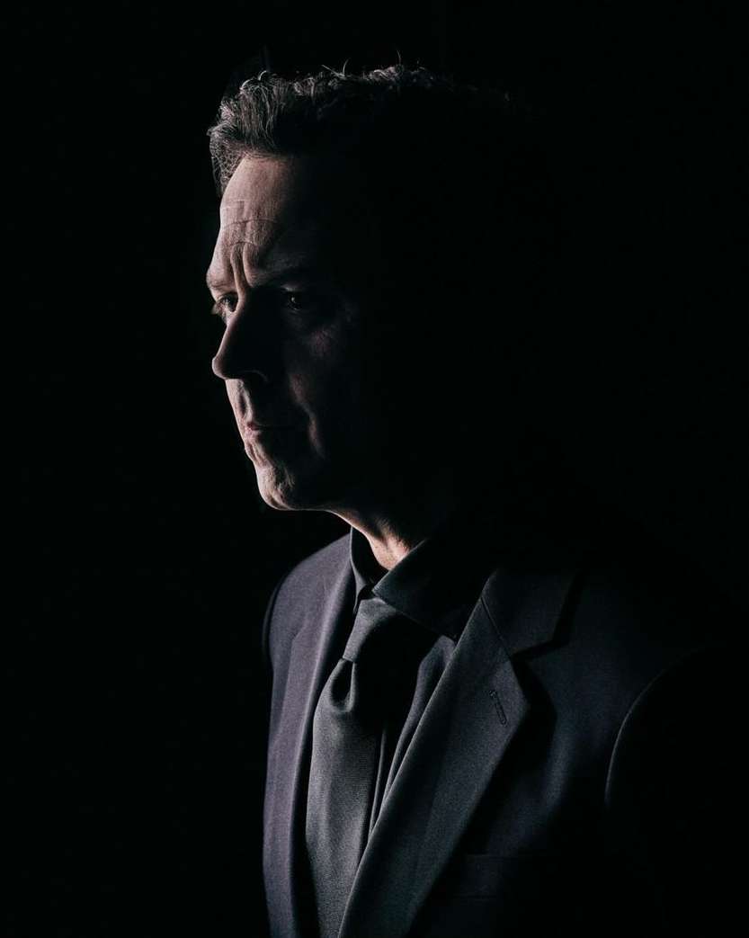 Portrait of a middle-aged man in a suit and tie, his face partially lit against a dark background, evoking a sense of mystery and formality.