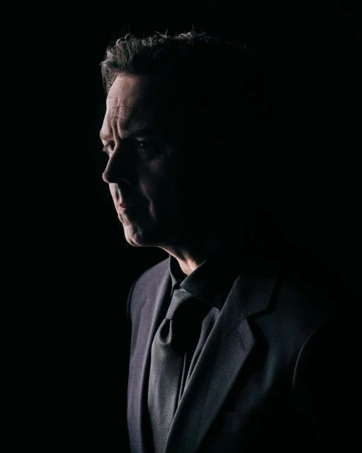 Portrait of a middle-aged man in a suit and tie, his face partially lit against a dark background, evoking a sense of mystery and formality.