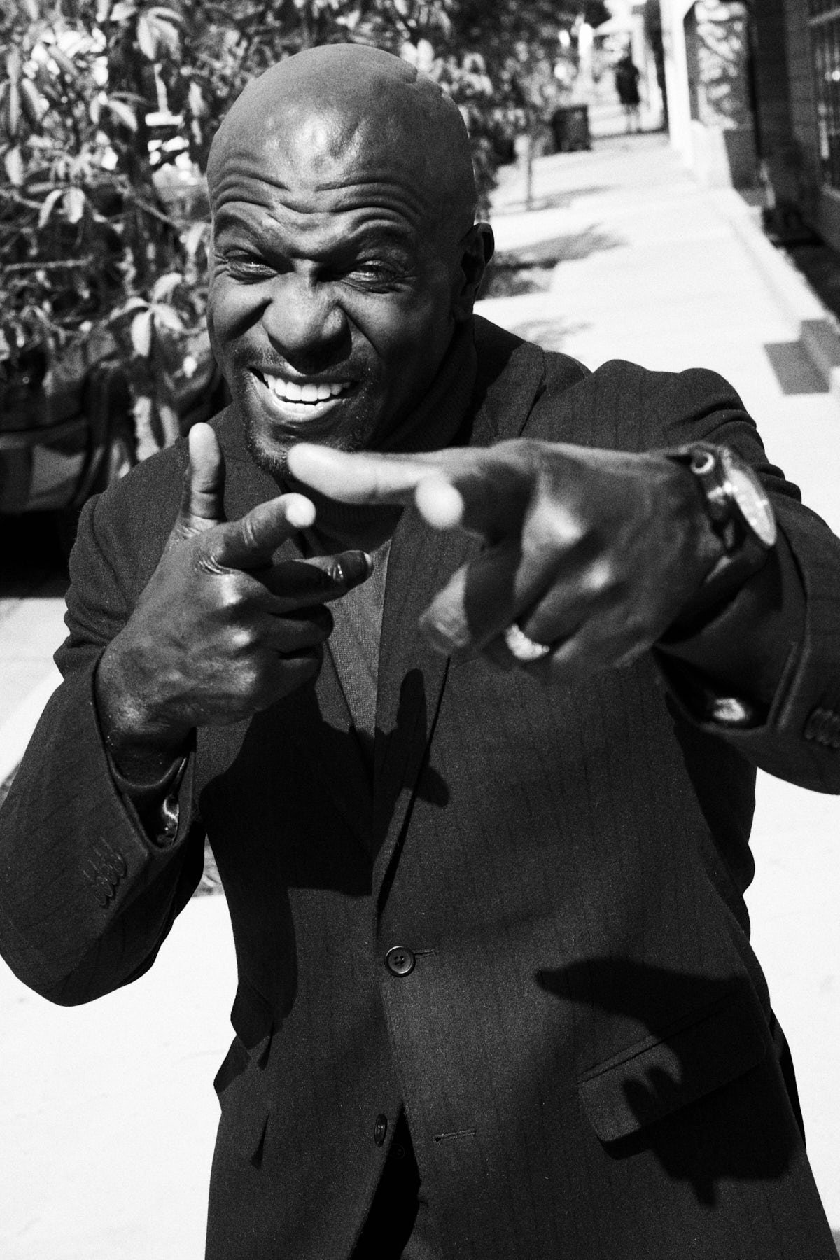 Monochrome portrait of an African American man in a suit pointing with finger guns against an outdoor setting with trees and a building.