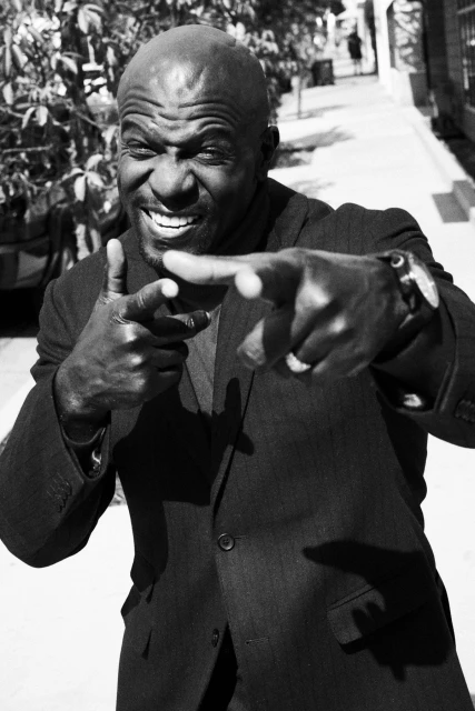 Monochrome portrait of an African American man in a suit pointing with finger guns against an outdoor setting with trees and a building.