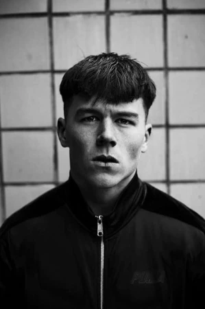 Close up portrait of a young man with dark hair wearing a zip up jacket against a tiled wall background