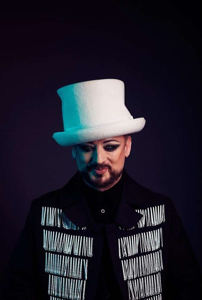 Portrait of a person with a beard wearing a white top hat and a black jacket with fringe, set against a dark purple background.