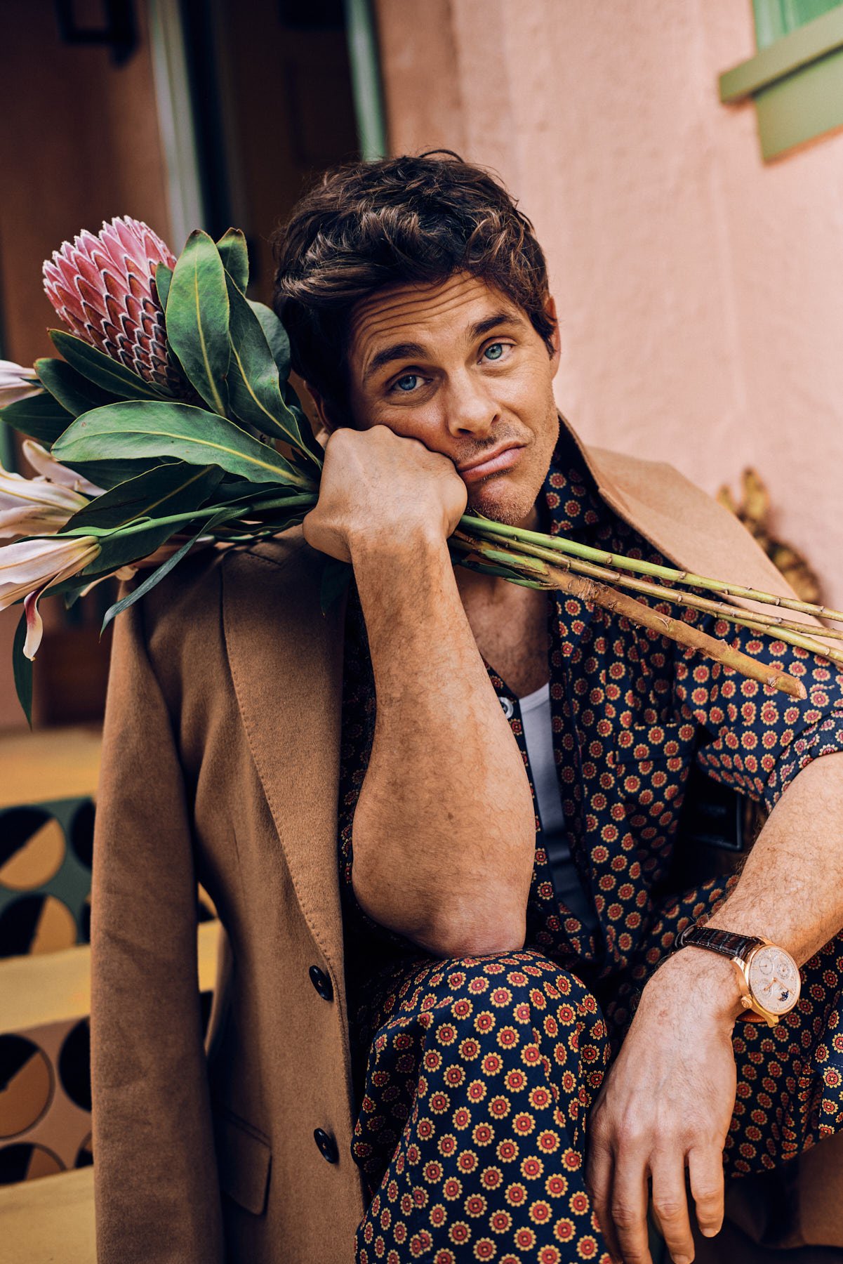 Adult male model in patterned shirt and suit jacket poses with flowers outdoors, resting his cheek on his hand in an outdoor setting.