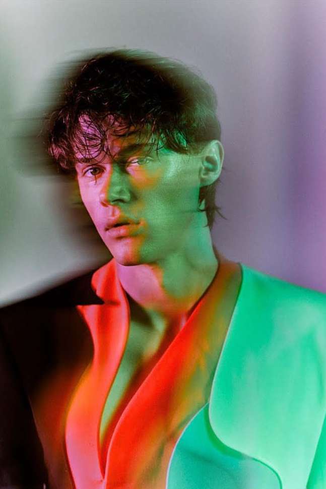 Portrait of a young man in a light colored blazer with a shadowed face and vibrant multi-colored studio lighting.