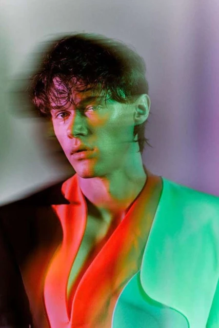 Portrait of a young man in a light colored blazer with a shadowed face and vibrant multi-colored studio lighting.