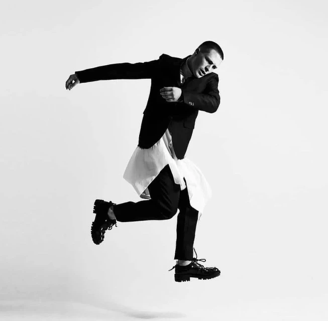A male fashion model in a black suit jacket and trousers is leaping mid-air in a studio with a light gray background, monochrome.