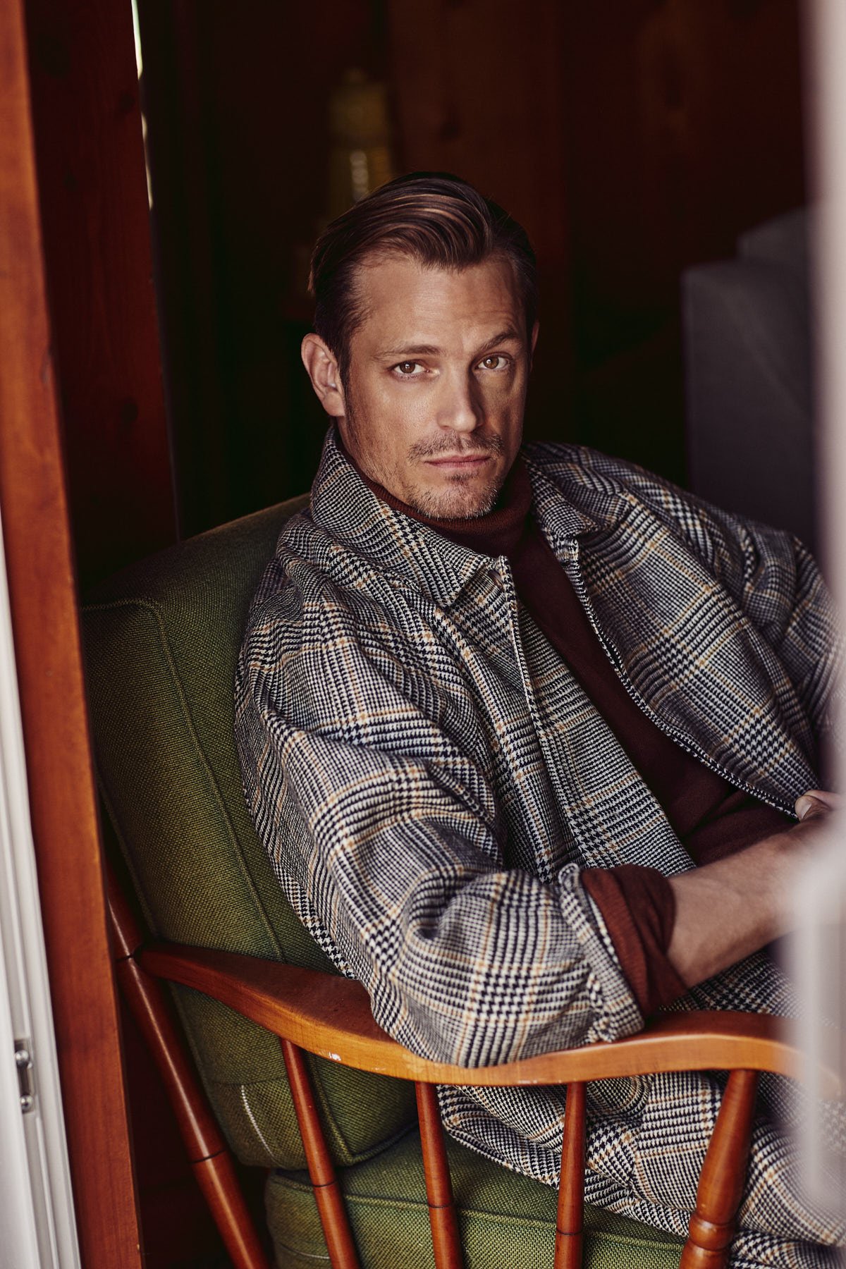 Portrait of Alexander Skarsgård in a plaid jacket and maroon turtleneck, sitting in a green upholstered chair inside a wood-paneled room.