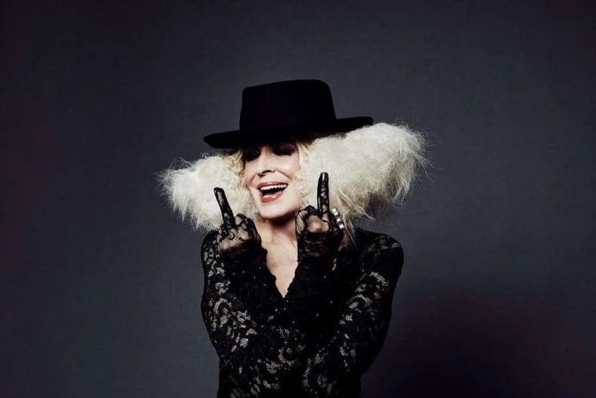 Elderly woman wearing a black hat, lace top, and gloves, making a hand gesture, photographed by Leigh Keily.