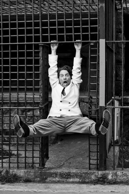 Monochrome portrait of a man in a light-colored suit hanging from metal bars with a surprised expression and outstretched legs.