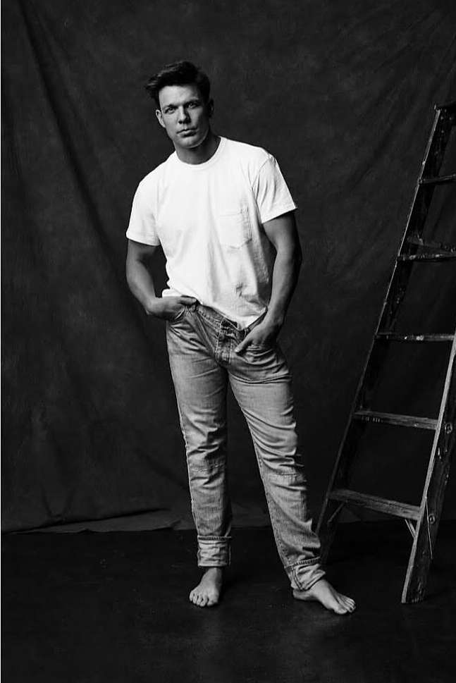 Barefoot adult male in white t-shirt and jeans poses in a photo studio near a wooden ladder against a dark mottled backdrop.