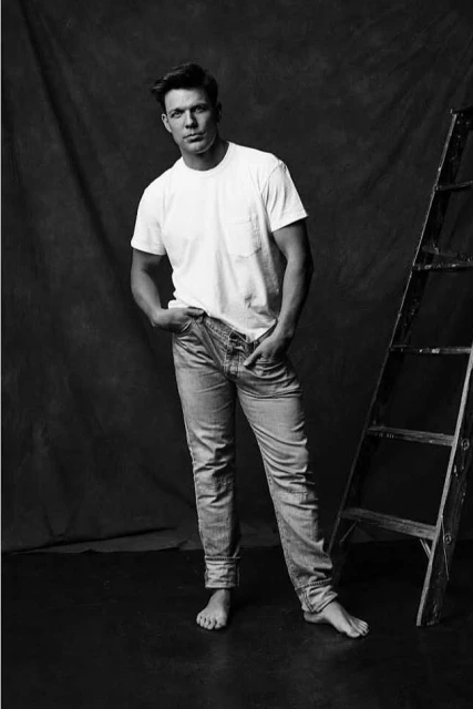 Barefoot adult male in white t-shirt and jeans poses in a photo studio near a wooden ladder against a dark mottled backdrop.
